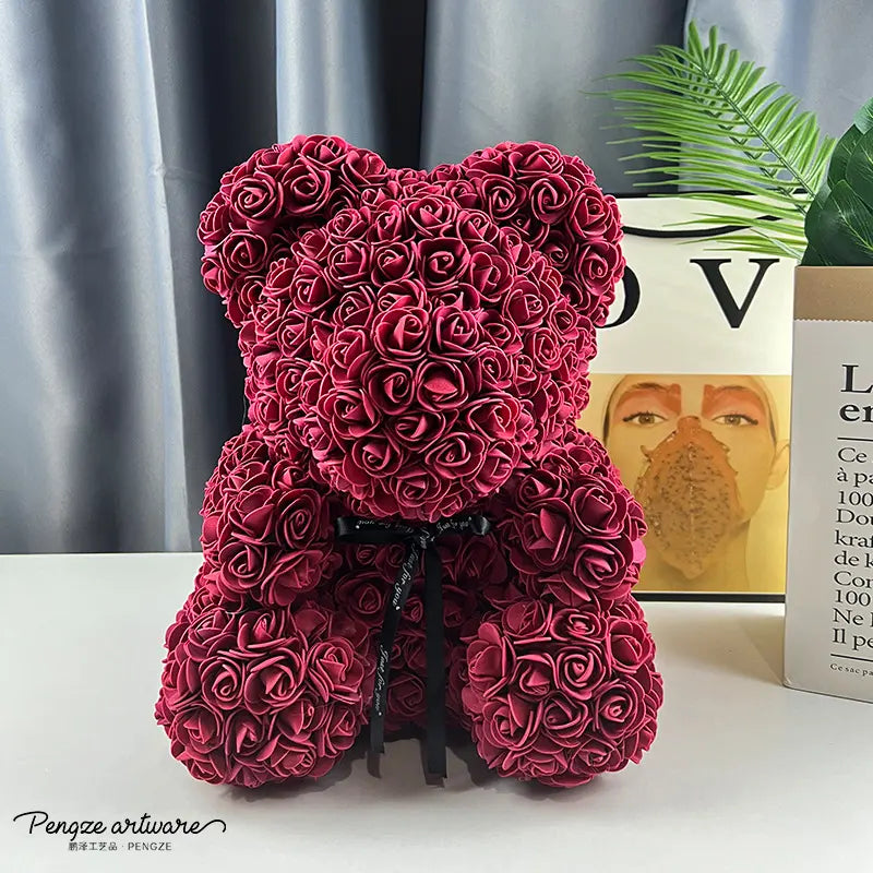 Plush teddy bear made of red roses, sitting on a white surface, with books and plant in background.