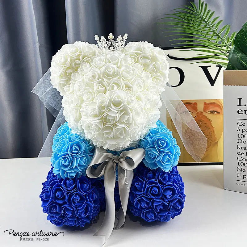 Bear-shaped rose arrangement in white and blue hues, with a silver crown and bowtie, on a table.