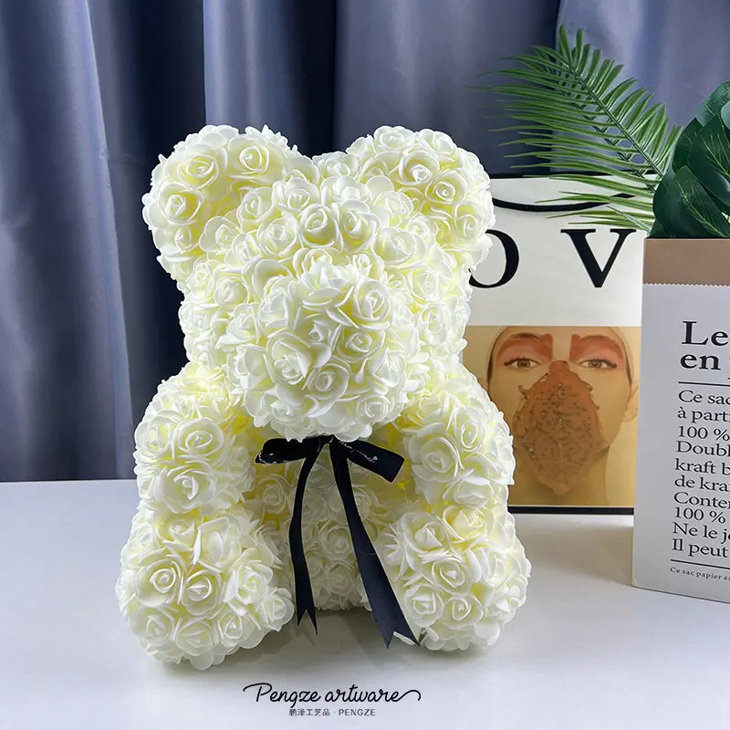 Teddy bear made of white roses with a black ribbon, set on a table with books and a plant.