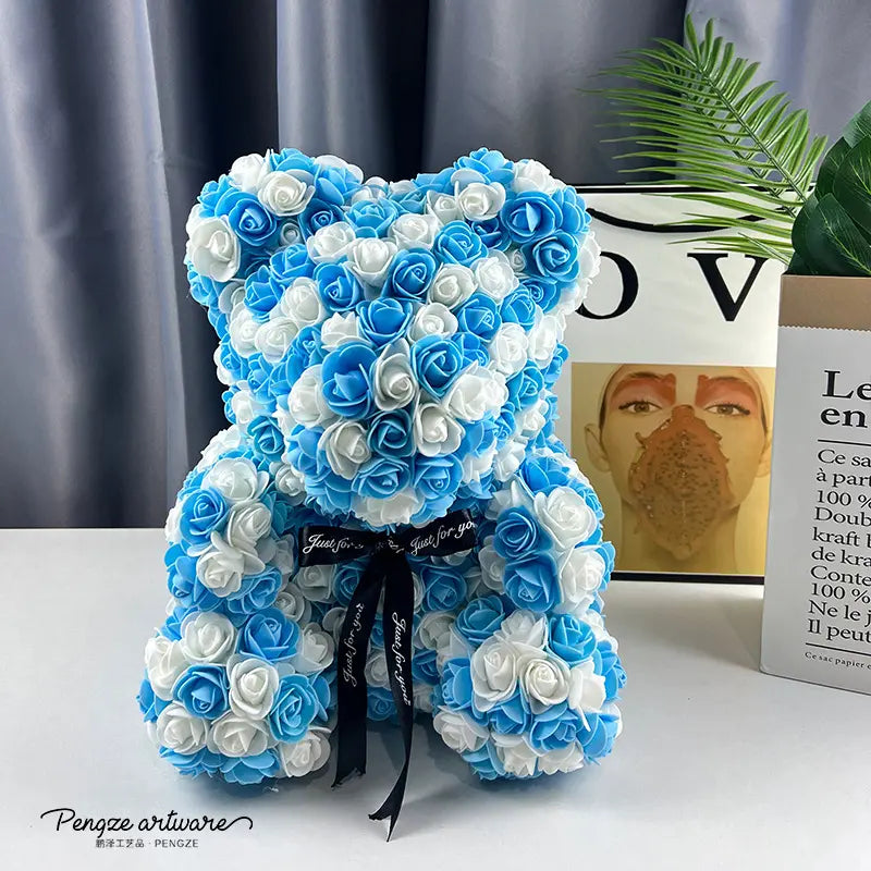 Teddy bear made of blue and white roses with a black ribbon, on a table with books and a plant.