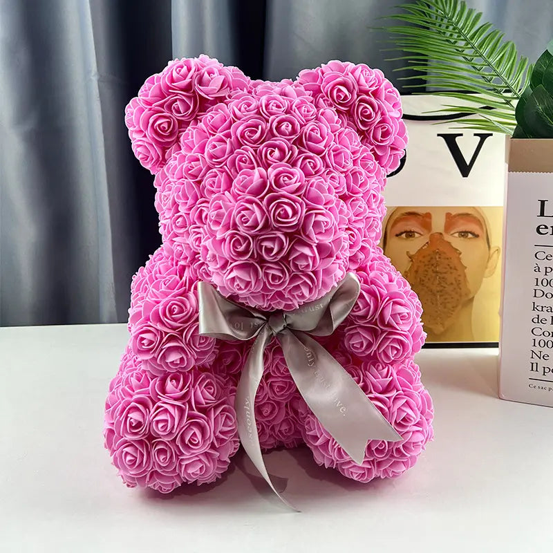 Pink teddy bear made of roses with a gray ribbon, set on a white table with a plant nearby.