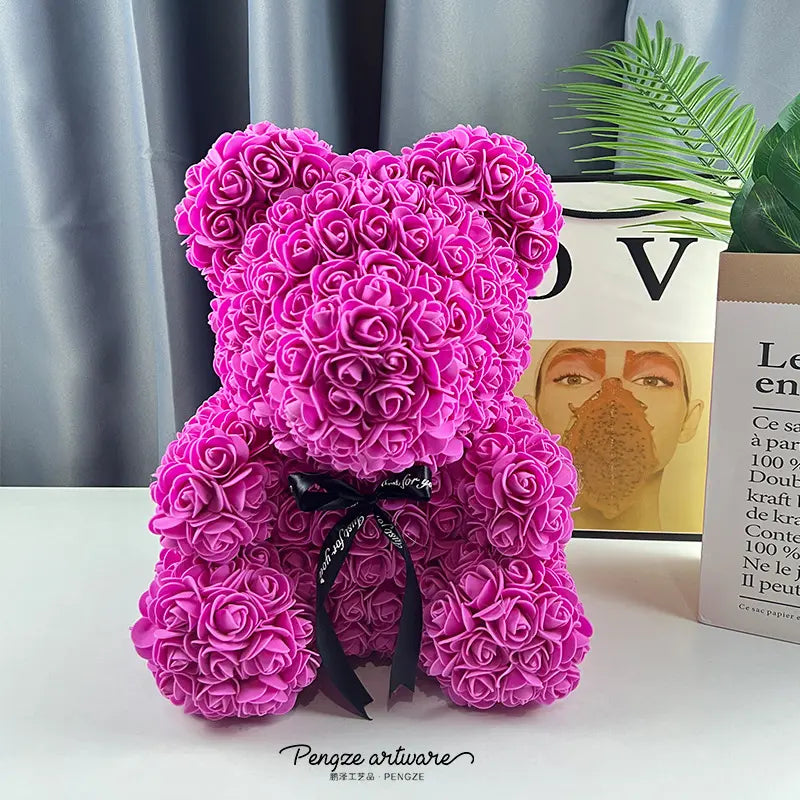 Pink rose teddy bear with a black ribbon sits on a white surface; gray curtain and plants behind.