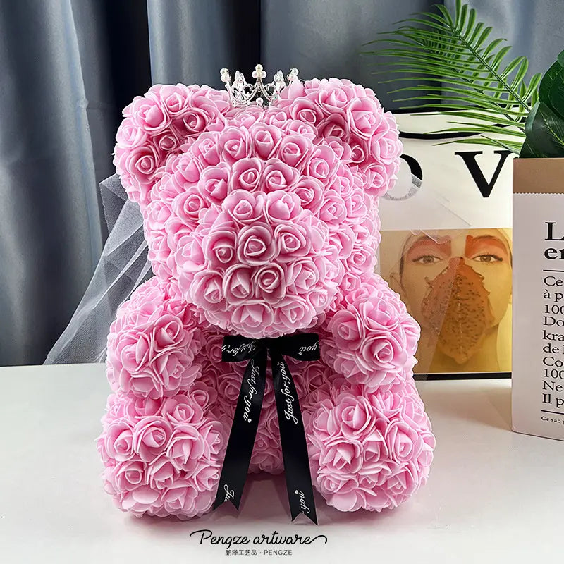 Pink rose teddy bear with a crown, black bow tie, and green leaves in the background.