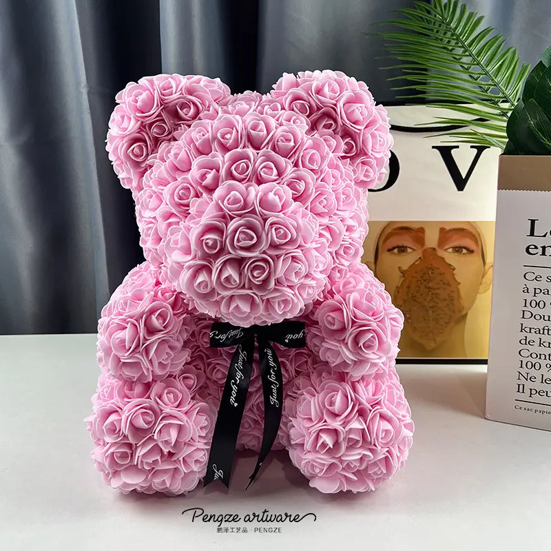 Pink teddy bear made of roses with a black ribbon, set against a book and a green plant.