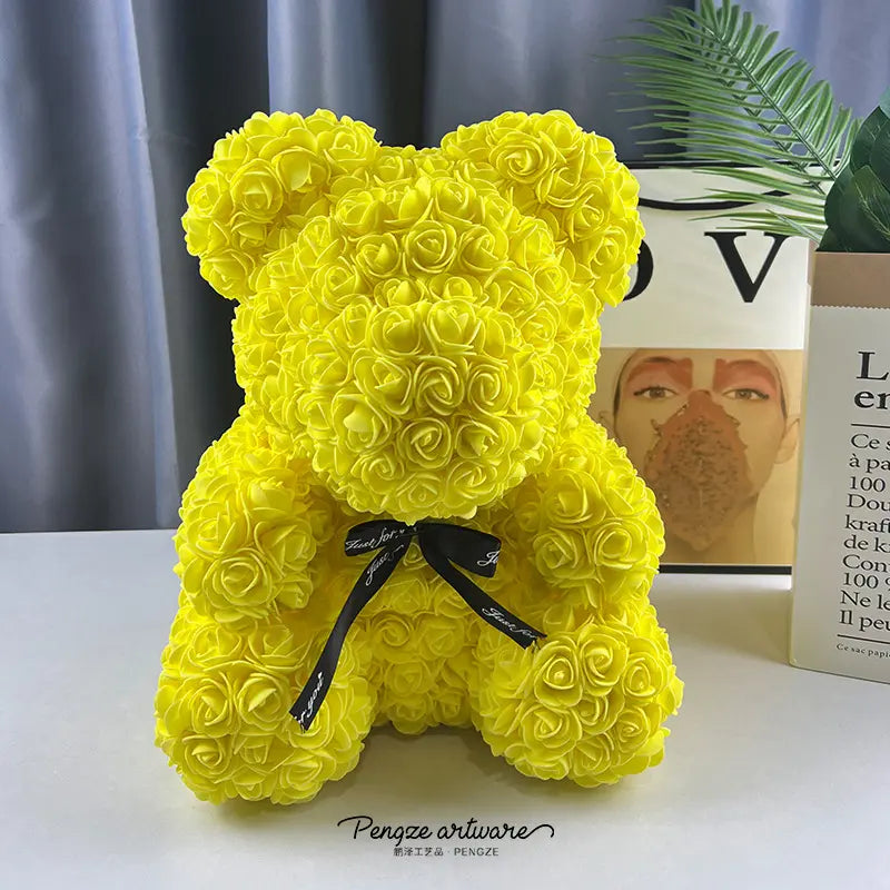 Yellow rose-covered teddy bear with a black bow, set against a gray curtain and plant backdrop.