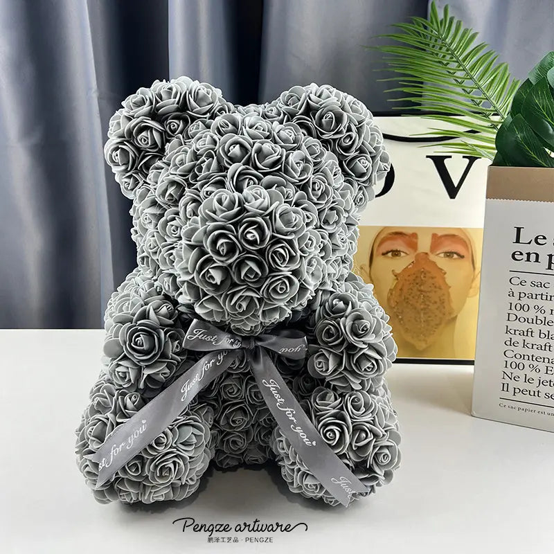 Gray rose teddy bear with ribbon sits on a white surface; green plant and books in background.
