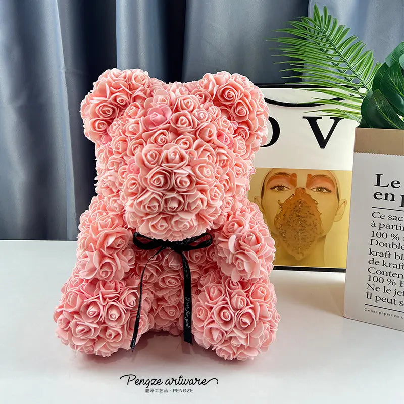 Pink rose-covered teddy bear with a black ribbon sits on a table, with books and a plant behind.