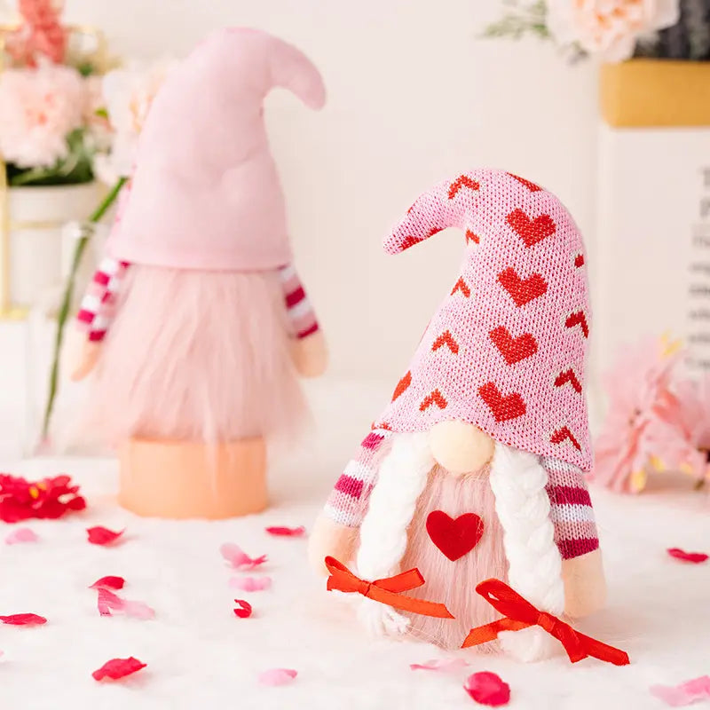 Two plush gnomes with pink hats and hearts, surrounded by soft pink petals and flowers.