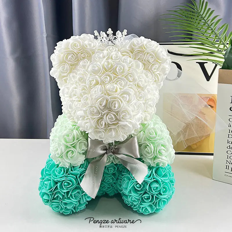 A teddy bear made of white, mint, and teal roses, with a silver bow and crown, on a table.
