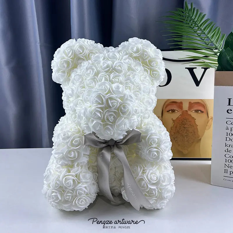 White teddy bear made of roses with a gray bow, set against a blue curtain and framed artwork.