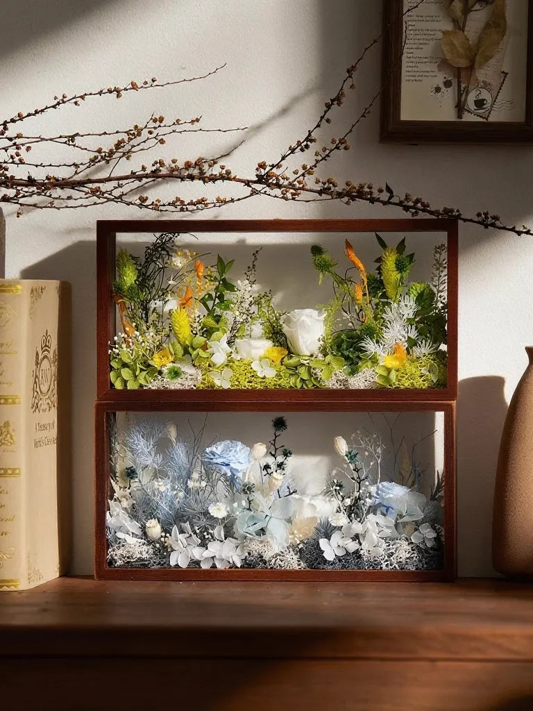 Two wooden-framed boxes with colorful dried flowers on a wooden shelf, sunlight casting shadows.