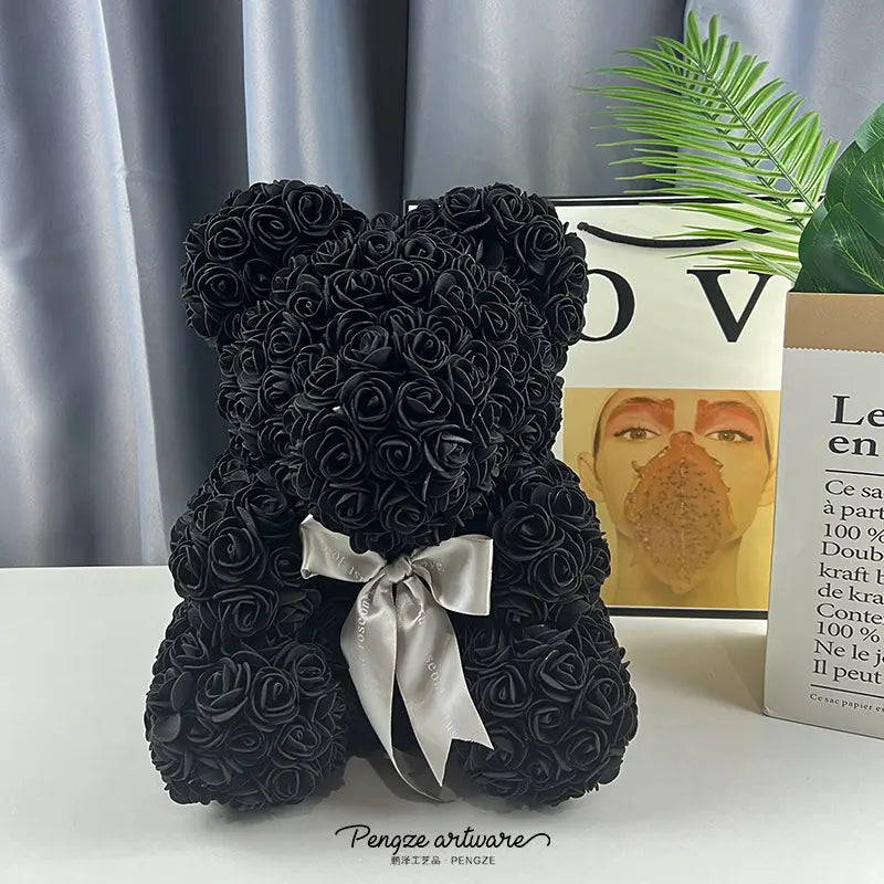 Black rose teddy bear with a white bow, set against books and a plant on a white surface.