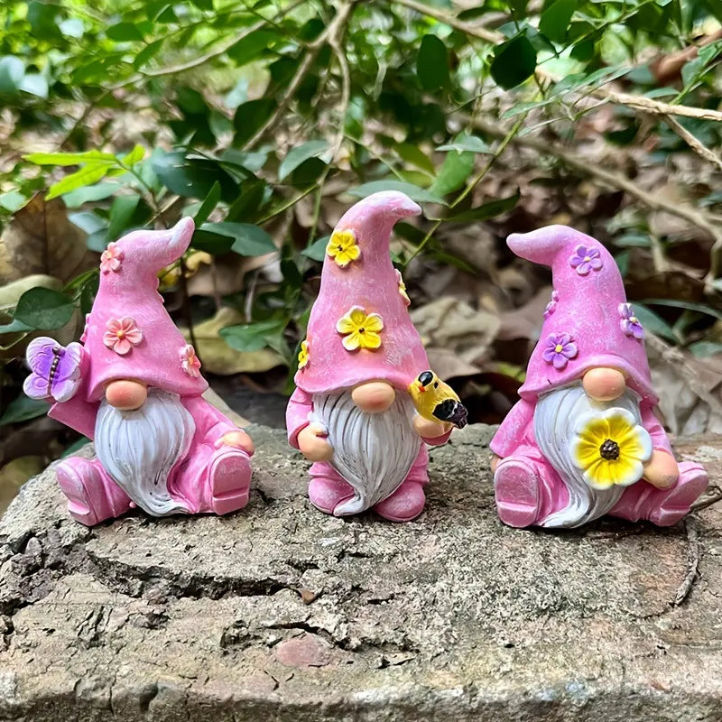 Three pink gnome figurines with flowers sit on a rock, surrounded by greenery.