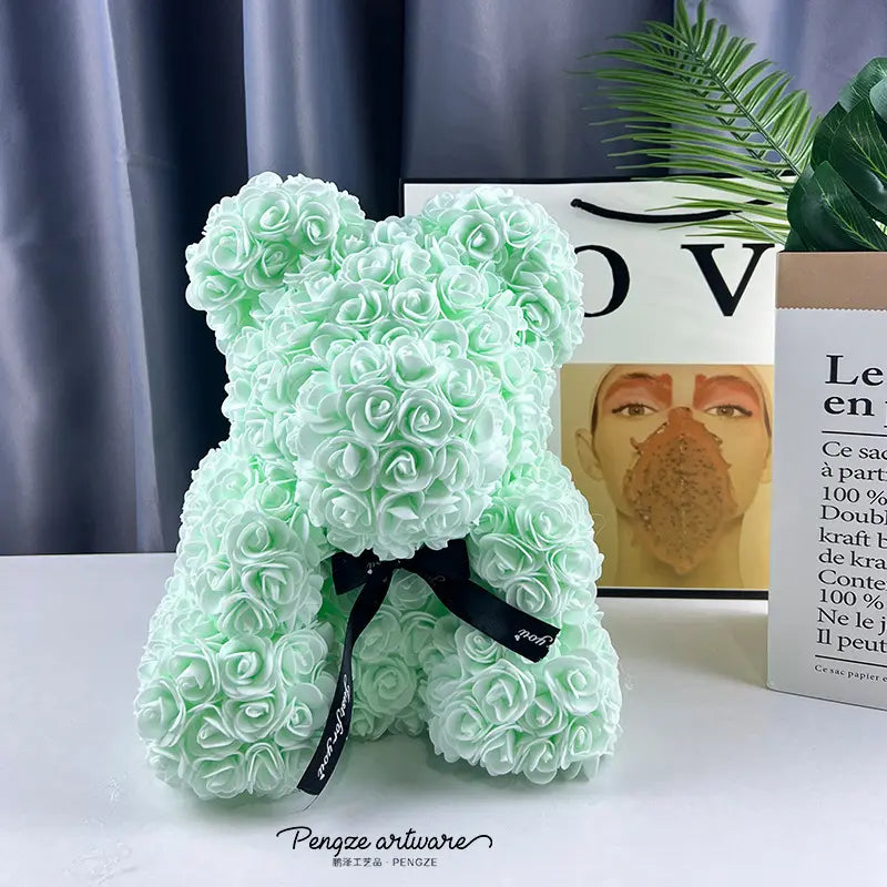 Mint green rose teddy bear with a black bow, set against books and a plant on a white surface.
