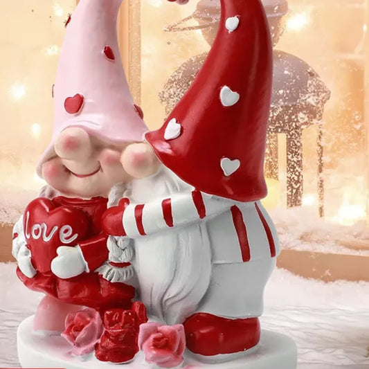 Two gnomes in red and pink hats with heart patterns hug in a snowy, lantern-lit scene.