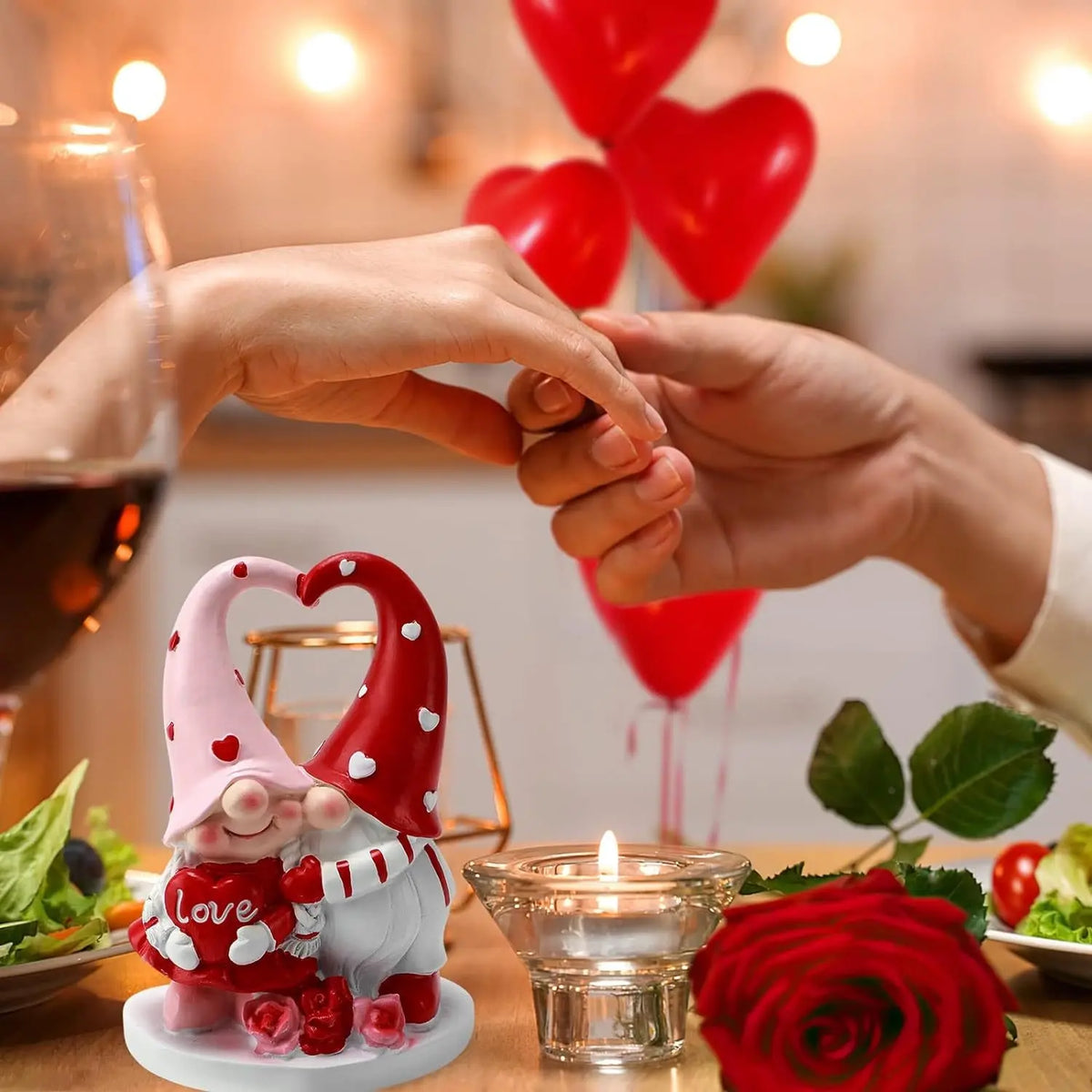 Two hands holding over a table with a heart-themed gnome, red balloons, wine, and a candle.