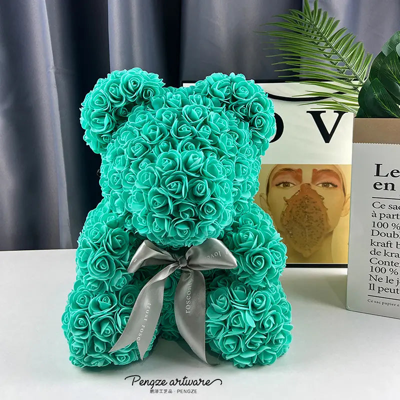 A teal rose-covered teddy bear with a silver bow, on a table against a gray curtain and plant.