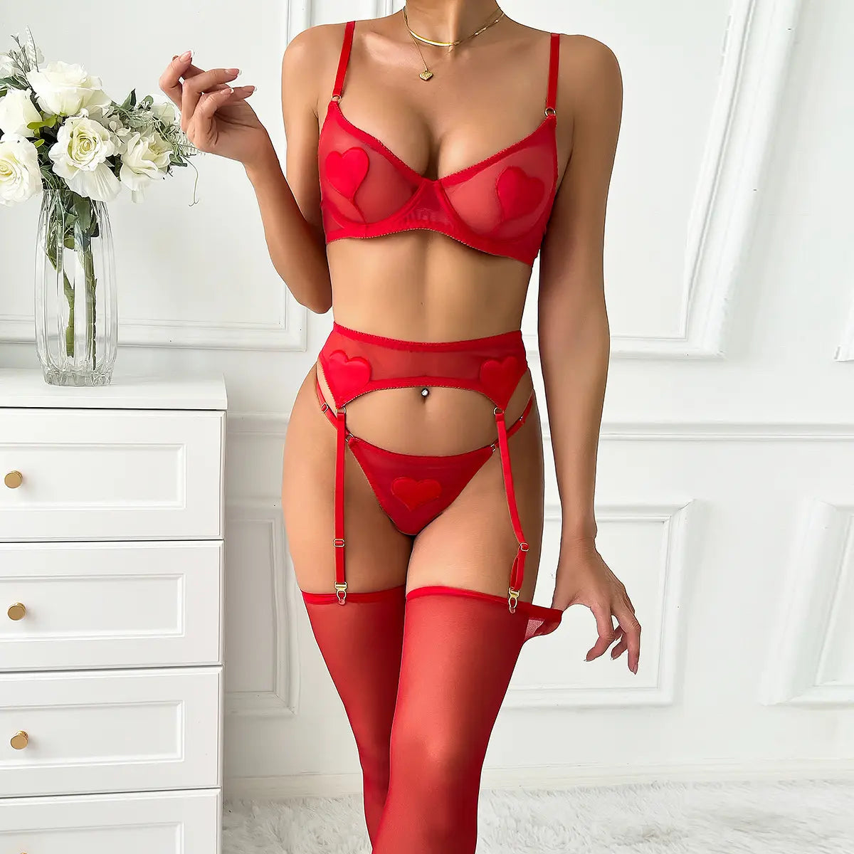 A woman wears red lingerie with heart details, a garter belt, and stockings in a white room.