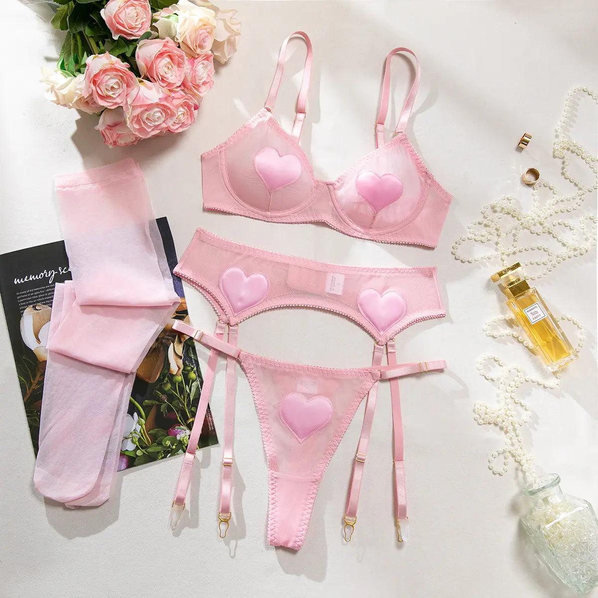 Pink lingerie set with heart designs, garter belt, stockings, roses, and a perfume bottle nearby.