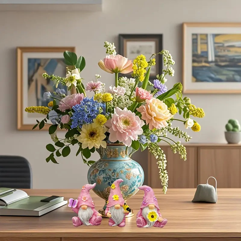 Vase with colorful flowers and three pink gnomes on a wooden desk, framed art in background.
