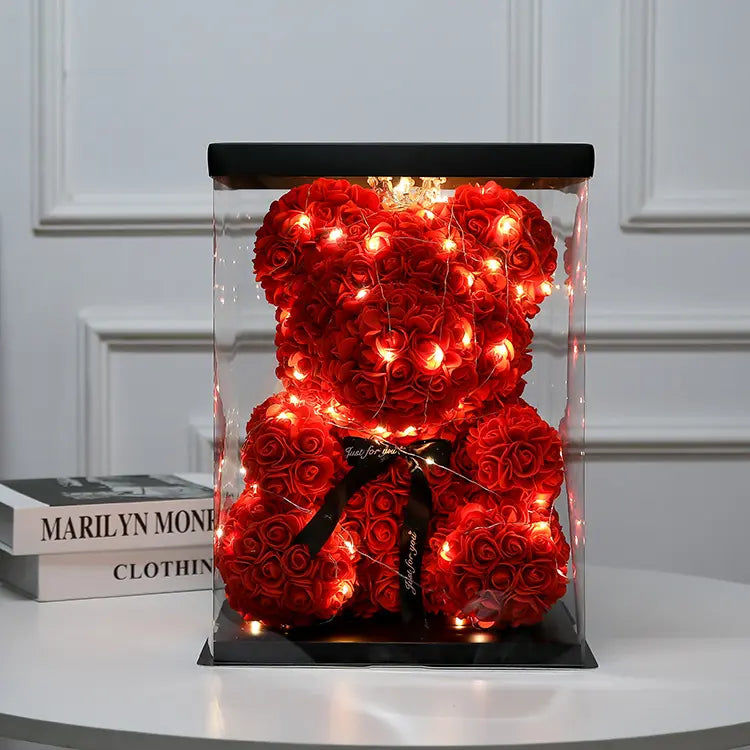 Red rose teddy bear with fairy lights in a clear box, on a white table beside stacked books.