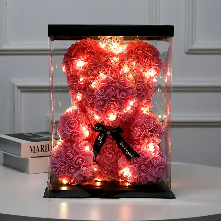 Pink rose teddy bear with string lights in a clear box, black ribbon, on a white table.